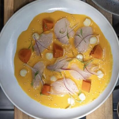 Fatias finas de peixe branco em leite de tigre amarelo, com batata-doce e milho, num prato visto de cima.