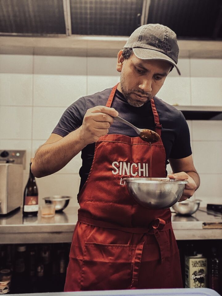 O chef Joseph Rios Aban na cozinha, de boné e avental vermelho com o nome Sinchi, com uma taça de inox numa mão e uma colher na outra.