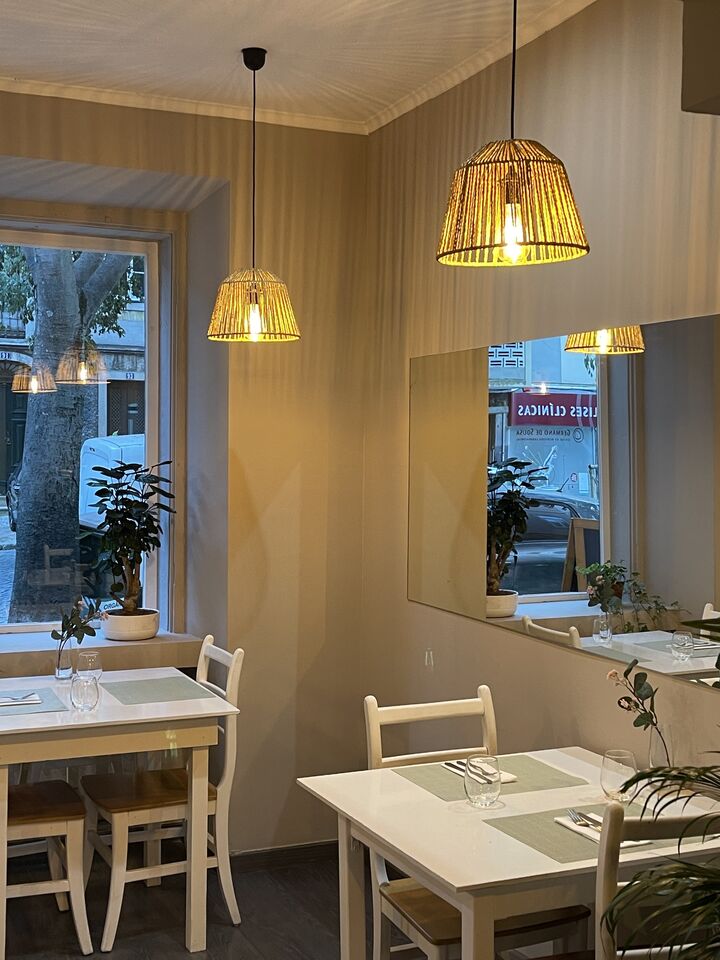 A sala de dia: mesas brancas junto à janela da rua, candeeiros de vime e um espelho grande na parede.
